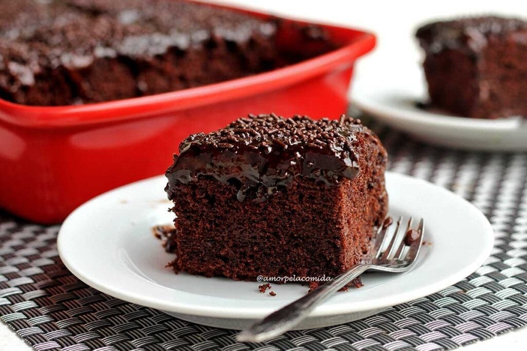 Fatia retangular de bolo de chocolate com cobertura de chocolate sobre prato redondo branco, ao fundo a forma que foi assado o bolo