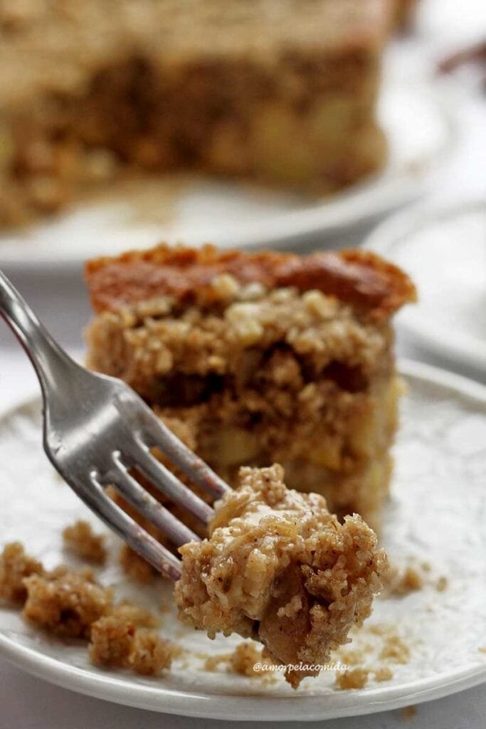 Garfo pequeno com um pedacinho de torta de maçã. ao fundo o pedaço dentro de um prato redondo branco