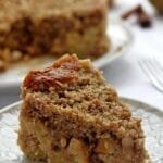 Fatia de torta de maçã com cobertura de farofa crocante sobre prato redondo, ao fundo a torta de maçã partida