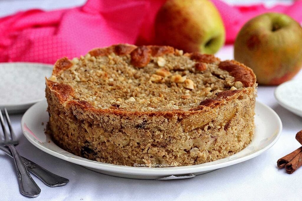 Torta de maçã redonda sobre prato redondo branco, ao fundo duas maçãs, ao lado dois garfinhos