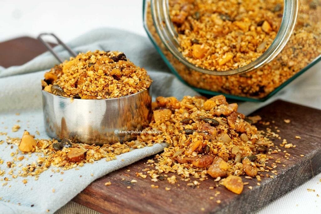 Granola salgada dentro de uma xícara de metal, ao lado um pouco sobre uma tábua de madeira e ao fundo o vidro deitado com a granola dentro