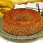 Bolo redondo com furo no meio sobre prato verde com detalhes na cor creme, ao fundo um cacho de banana e alguns limões
