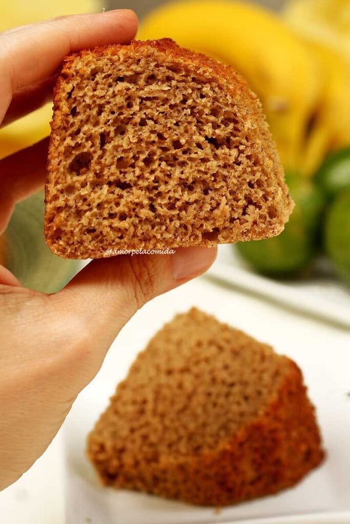 Mão segurando fatia de bolo inteiro, abaixo uma fatia de bolo deitada sobre um prato quadrado branco, pequeno