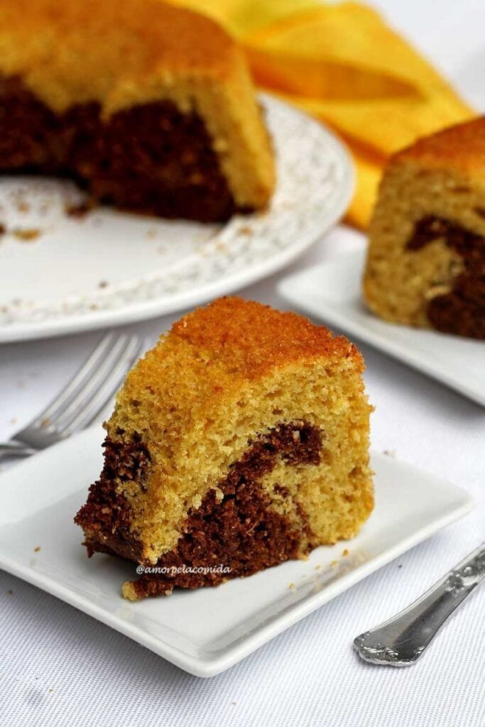 Duas fatias de bolo de fubá com uma parte mesclada de chocolate, os pedaços estão sobre 2 pratos pequenos quadrados