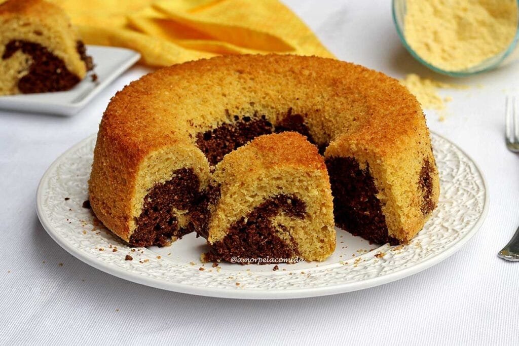 Bolo redondo com furo no meio de fubá, a massa é mesclada com chocolate, na parte inferior do bolo percebemos a maior concentração da massa de chocolate e na parte superior o bolo mais amarelinho de fubá. O bolo está cortado, na parte central tem uma fatia de bolo onde a parte mesclada de chocolate parece uma onda