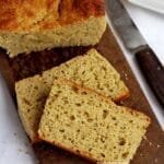 Duas fatias de pão sobrepostas sobre uma tábua de madeira retangular de cor escura, ao lado uma faca e o pão que foi cortado as fatias