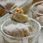 Colher pequena servindo uma porção de sobremesa de pêssego, abaixo o potinho da sobremesa, ao fundo uma parte da travessa